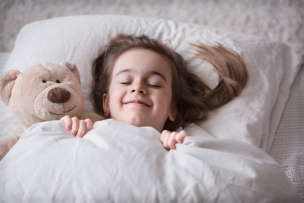 Cute little girl sleeps sweetly in a white cozy bed with a soft bear toy, the concept of children's rest and sleep